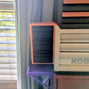 May include: An orange and black storage cube sits on a purple shelf. The cube has a rectangular shape and is open in the front, revealing black contents. A white object with the word "KOB" is to the right.