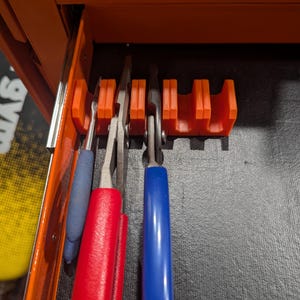 May include: An orange plastic tool organizer rack with multiple slots, mounted inside an orange metal drawer labeled RATCHETS. The rack holds three metal hand tools with blue and red rubberized handles, positioned vertically in the storage slots.