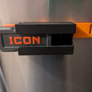 May include: A black and orange tool holder with the word "ICON" in orange lettering. The holder is mounted on a metallic surface. The holder has a rectangular shape with a slot for a tool and an orange extension.