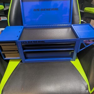 May include: A blue US GENERAL toolbox with multiple drawers and compartments. The toolbox is open, revealing a black interior and the US GENERAL logo. It is sitting on a black and green chair.
