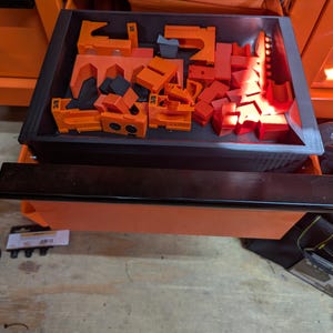 May include: A drawer filled with various orange and black tools. The tools are of different shapes and sizes, including clamps and blocks. The drawer is inside an orange cabinet, with the tools neatly organized within a black tray.