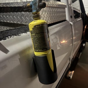 May include: A yellow propane torch with a blue and brass head is held in a black holder attached to a white truck. The torch has a black nozzle and is mounted on the side of the truck bed.