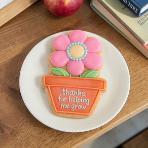 May include: A flower-shaped cookie with pink petals, a yellow center, and a turquoise border, sits atop an orange flower pot. The pot has the text "thanks for helping me grow" in white. The cookie is on a white plate.