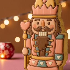 May include: A detailed cookie in the shape of a nutcracker soldier. The cookie features a pink, brown, and yellow color scheme with heart and crown details. The nutcracker holds a heart-shaped scepter. The background is blurred with warm tones.