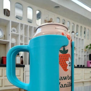 May include: A light blue 3D-printed can holder with a handle, holding a can of strawberry vanilla soda. The can features a strawberry graphic and the text "Strawberry Vanilla". The holder is on a gray surface.