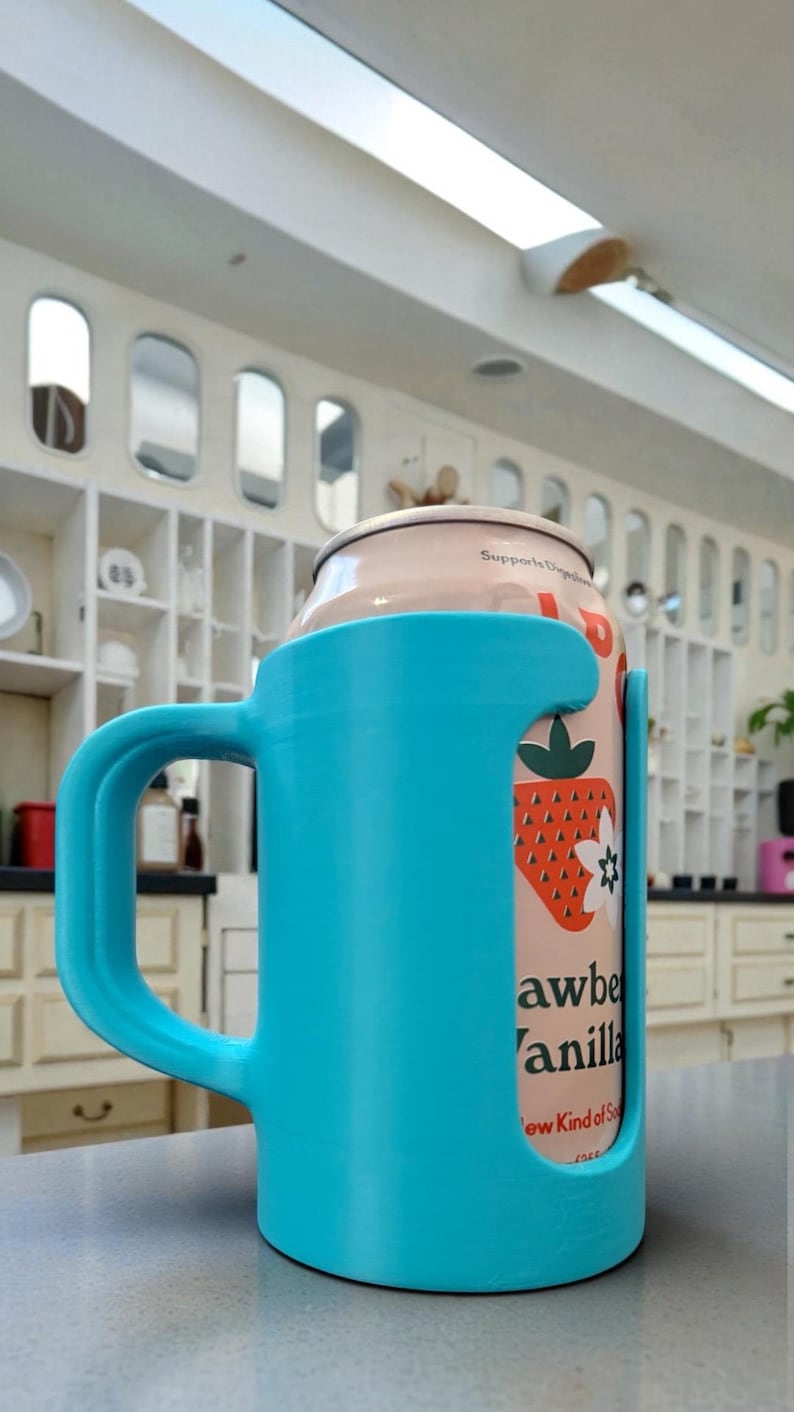 May include: A light blue 3D-printed can holder with a handle, holding a can of strawberry vanilla soda. The can features a strawberry graphic and the text "Strawberry Vanilla". The holder is on a gray surface.