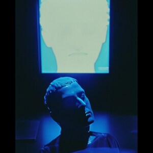 May include: A man's head is illuminated in blue light, looking up at a television screen with a blue and white image of a face. The text "The Fool" is displayed above the image.