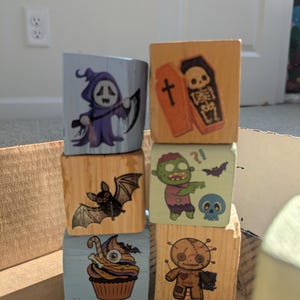 May include: Six wooden blocks stacked in two columns, featuring Halloween-themed illustrations including a purple ghost with a scythe, a skeleton in a coffin, a black bat, a green zombie, a skull, a decorated cupcake, and a brown voodoo doll.
