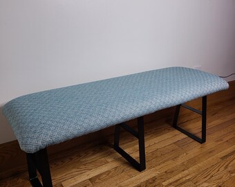 May include: A rectangular bench with a light blue patterned cushion and black metal legs. The bench is approximately 48 inches long and is placed against a white wall and wooden floor.