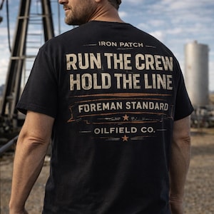 Może przedstawiać: Czarna koszulka z napisem "RUN THE CREW HOLD THE LINE FOREMAN STANDARD OILFIELD CO." w postarzanym białym i pomarańczowym napisie. Koszulka jest noszona przez osobę noszącą biały kask i okulary przeciwsłoneczne.