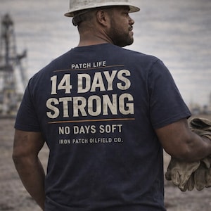 Może przedstawiać: Granatowa koszulka z nadrukiem "14 DAYS STRONG" i "NO DAYS SOFT" na plecach. Koszulka ma również napis "IRON PATCH OILFIELD CO.". Osoba ma na sobie biały kask i trzyma rękawice robocze.