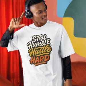Op de afbeelding: Wit T-shirt met de tekst "Stay Humble Hustle Hard" in een gestileerd ontwerp. Het shirt heeft een ronde hals en korte mouwen. De persoon op de afbeelding draagt een zwarte koptelefoon en zwarte mesh mouwen.