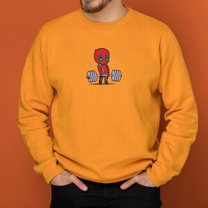 May include: A yellow sweatshirt with a cartoon image of Deadpool lifting a barbell. The image is in the center of the sweatshirt.