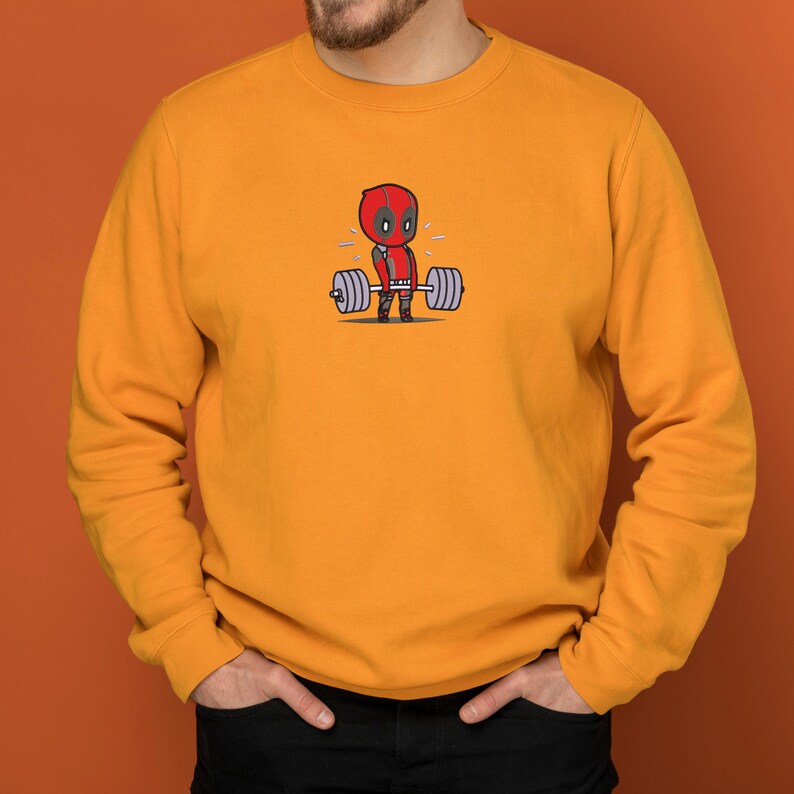 May include: A yellow sweatshirt with a cartoon image of Deadpool lifting a barbell. The image is in the center of the sweatshirt.