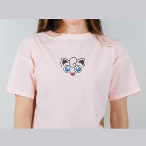 May include: A pink t-shirt with a cartoon image of a pink Pokemon character with blue eyes and a happy expression.