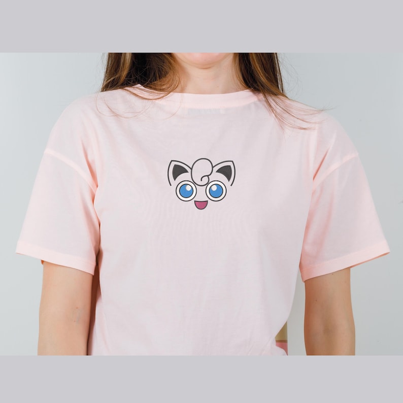 May include: A pink t-shirt with a cartoon image of a pink Pokemon character with blue eyes and a happy expression.