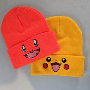May include: Two knit beanies with embroidered cartoon faces. The beanie on the left is red with a red, blue, and white cartoon face. The beanie on the right is yellow with a yellow, red, and white cartoon face.