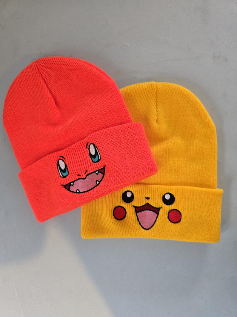 May include: Two knit beanies with embroidered cartoon faces. The beanie on the left is red with a red, blue, and white cartoon face. The beanie on the right is yellow with a yellow, red, and white cartoon face.
