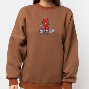 May include: A brown sweatshirt with a red and black cartoon image of Deadpool lifting a barbell. The sweatshirt has a red ribbed collar and cuffs.