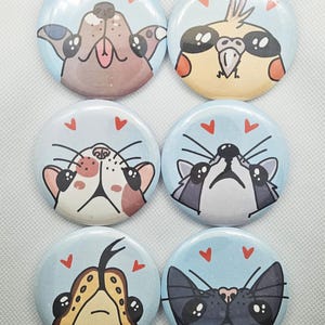 May include: Six circular pin badges with cartoon animal faces. The badges have a light blue background and red heart details. The animal faces include a dog, bird, cat, and raccoon, each with a unique expression.
