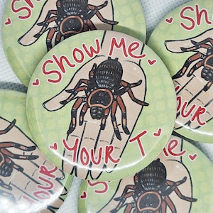 Op de afbeelding: Een verzameling groene, ronde knopen met een handgetekende illustratie van een spin op een hand. De tekst "Show Me Your T's" is rood. De spin is zwart en bruin.
