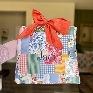May include: A patchwork tote bag with a large orange bow. The bag features a variety of floral and patterned fabrics in red, blue, yellow, and green. The name "Jaci" is embroidered in blue thread on the front.