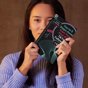 May include: A person holding a black and teal booklet with the text "You're 5 Steps Away from Your First 10K TikTok Follow" and "2025". The booklet also has the words "Instant Download" and "LI". The person is wearing a light blue jumper.