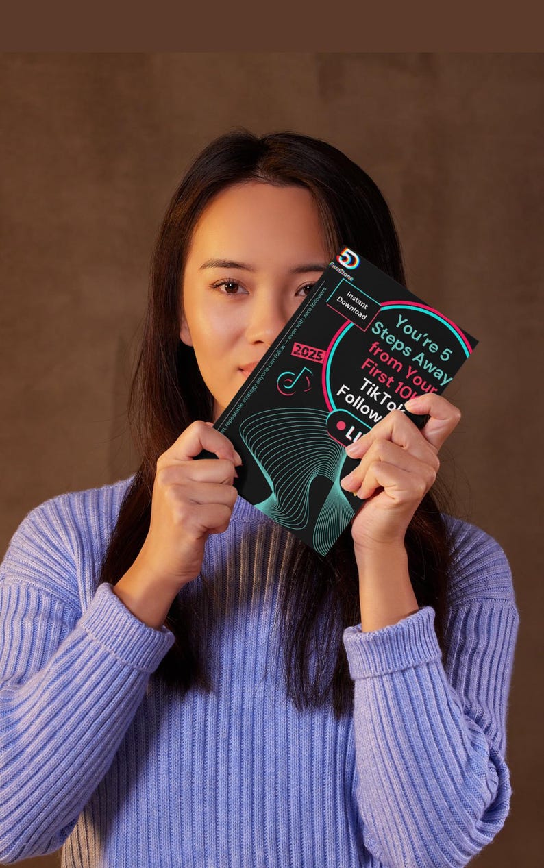 May include: A person holding a black and teal booklet with the text "You're 5 Steps Away from Your First 10K TikTok Follow" and "2025". The booklet also has the words "Instant Download" and "LI". The person is wearing a light blue jumper.