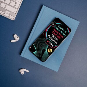 May include: A smartphone with the text "You're 5 Steps Away from Your First 10K TikTok Followers" on the screen, set on a blue book. A silver keyboard and white earbuds are on a blue surface. The phone has a black and teal design.