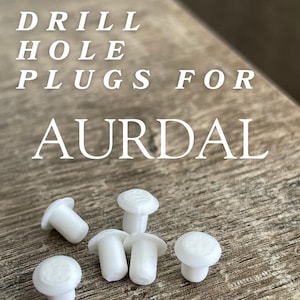 May include: White drill hole plugs for AURDAL furniture. The image shows several small, mushroom-shaped plugs with a flat, circular top and a cylindrical stem. The text in the image reads "DRILL HOLE PLUGS FOR AURDAL".