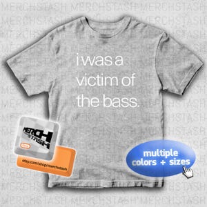 May include: A light gray t-shirt with the text "I was a victim of the bass." in white. The shirt has a crew neck and short sleeves. Additional graphics include a shop logo and a blue oval with the text "multiple colors + sizes."