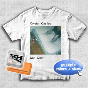 May include: White t-shirt featuring the text "Crystal Castles" and "Doe Deer." The shirt has a rectangular image of a person sitting on a mattress. The shirt also has a blue oval with the text "multiple colors + sizes."