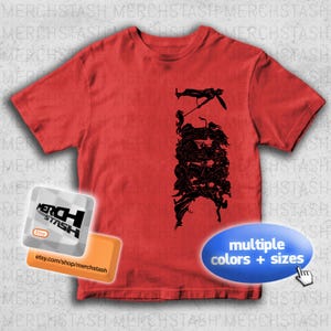 May include: A red t-shirt featuring a black graphic design of figures in action. The shirt has a crew neck and short sleeves. The image also includes a logo with the text "MERCHSTASH" and a callout for multiple colors and sizes.