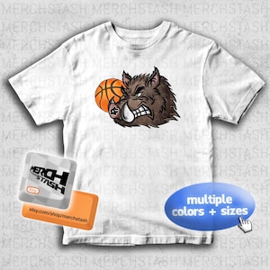 Könnte beinhalten: Weißes T-Shirt mit einer Cartoon-Illustration eines knurrenden Wildschweins, das einen Basketball hält. Das Wildschwein ist braun mit weißen Stoßzähnen und einem grimmigen Gesichtsausdruck. Der Basketball ist orange. Das Shirt hat auch den Text "multiple Farben + Größen".