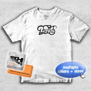 Puede incluir: Camiseta blanca con un diseño gráfico negro con los números "2025" y caras de dibujos animados. La camiseta tiene cuello redondo y mangas cortas. La imagen también incluye un logotipo de la tienda y una llamada para múltiples colores y tallas.