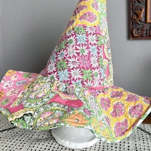 May include: A tall, pointed hat made of quilted fabric featuring a patchwork design. The pattern consists of various floral and paisley motifs in vibrant shades of pink, yellow, green, and white, with visible stitching throughout the fabric.