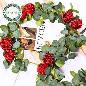 May include: A floral arrangement featuring red roses and green eucalyptus leaves. The garland is draped over a wooden structure, with a magazine visible in the background. The word "Wreathlyn" is in a circular logo.