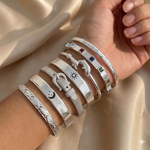Può includere: Una collezione di braccialetti d'argento con vari design, tra cui pesci, lune crescenti, soli e una fascia in stile cintura a rete. Alcuni braccialetti presentano pietre preziose colorate e dettagli incisi. I braccialetti sono indossati su un polso.