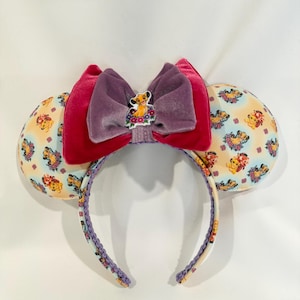 May include: A pair of mouse ears featuring a Simba theme. The ears are patterned with Simba and floral designs. A large, layered bow in shades of pink and purple sits atop the headband, with a Simba character embellishment.