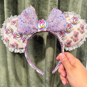 May include: A pair of mouse ears featuring a large, sequined lavender bow and Daisy Duck emblem. The ears are white with a Daisy Duck pattern and trimmed with white lace. The headband is held by a hand.