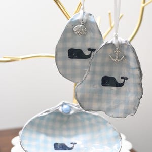 May include: Decorative seashell ornaments with a blue and white gingham pattern and a whale design. Each ornament is embellished with a small charm, including a seashell and an anchor. The ornaments are hanging from a gold-colored tree.