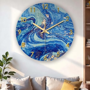 Puede incluir: Reloj de pared redondo con diseño de mármol azul y dorado. La esfera del reloj presenta números y manecillas doradas, con un patrón arremolinado azul y dorado. El reloj está montado en una pared blanca.