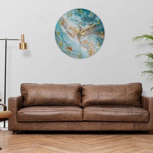 May include: Round wall clock with a teal and gold marble design. The clock features gold hands and gold numerals. The clock is hanging on a white wall above a brown leather sofa. Home decor.