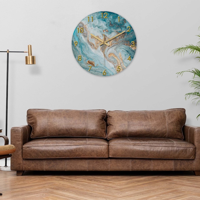 May include: Round wall clock with a teal and gold marble design. The clock features gold hands and gold numerals. The clock is hanging on a white wall above a brown leather sofa. Home decor.