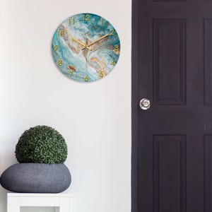 May include: Round wall clock with a teal and gold marble design. The clock features gold hands and gold numerals. The clock is hanging on a white wall. The clock is approximately 12 inches in diameter.