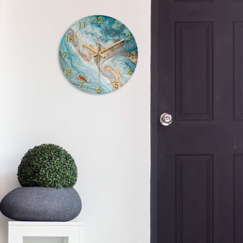 May include: Round wall clock with a teal and gold marble design. The clock features gold hands and gold numerals. The clock is hanging on a white wall. The clock is approximately 12 inches in diameter.