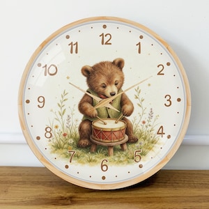 Puede incluir: Un reloj de pared redondo con marco de madera clara y esfera color crema. El reloj presenta números marrones del 1 al 12 y una ilustración de un oso pardo con chaleco verde, sentado en un taburete y tocando un tambor en un campo de hierba.