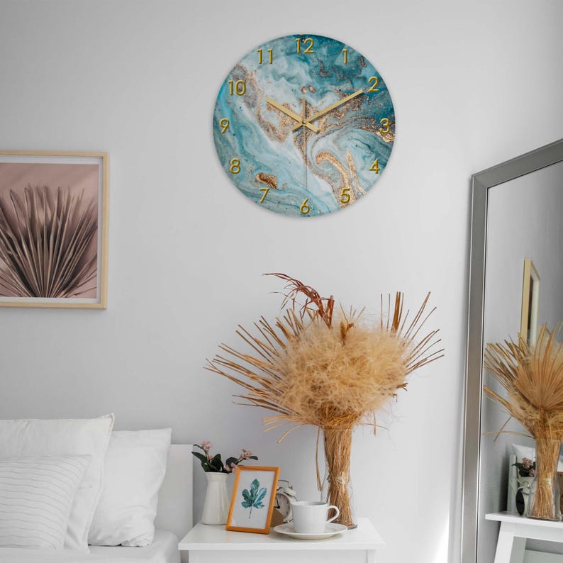 May include: Round wall clock with a teal and gold marble design. The clock features gold-colored numerals and hands. The clock is hanging on a white wall. The clock is a decorative item.