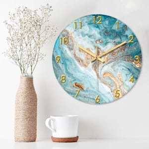 May include: Round wall clock with a teal and gold marble design. The clock features gold numerals and hands. A vase with white flowers and a white mug are also in the image.