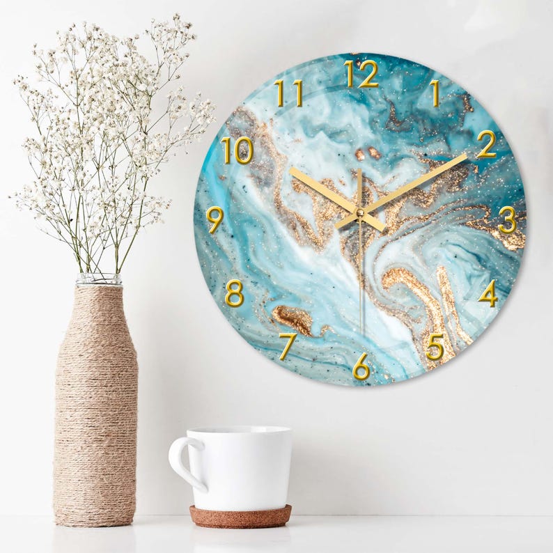 May include: Round wall clock with a teal and gold marble design. The clock features gold numerals and hands. A vase with white flowers and a white mug are also in the image.